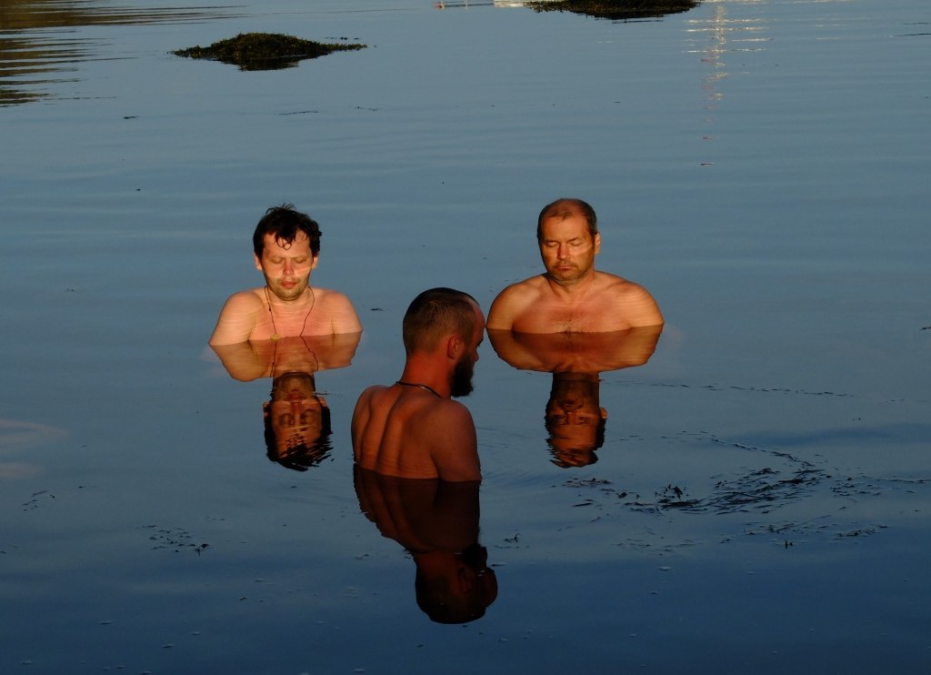 Три человека стоят по грудь в неподвижной воде Белого моря на закате, их отражения видны в водной глади